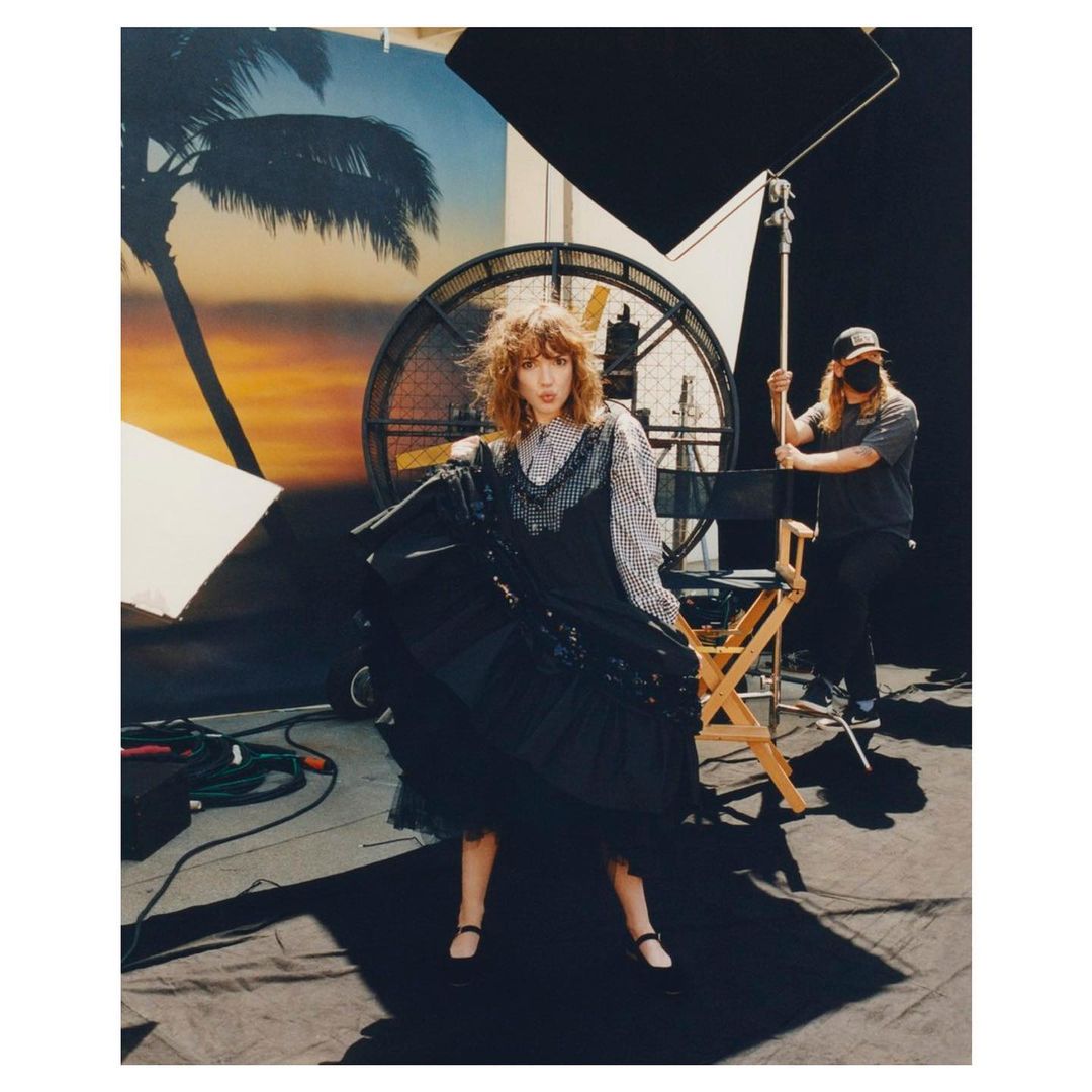 Fashion shot of a woman in a black dress on a set with a large fan and lighting equipment, attended by a masked technician.