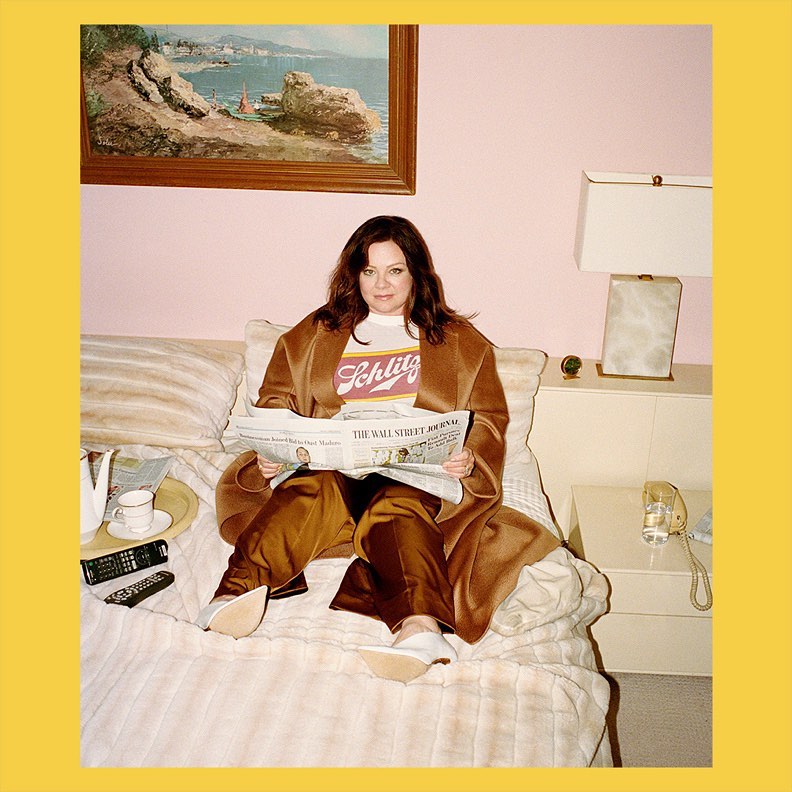 Melissa McCarthy seated on a bed reading a newspaper wearing a brown coat and white t-shirt