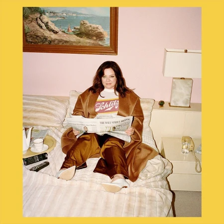 Melissa McCarthy seated on a bed reading a newspaper wearing a brown coat and white t-shirt