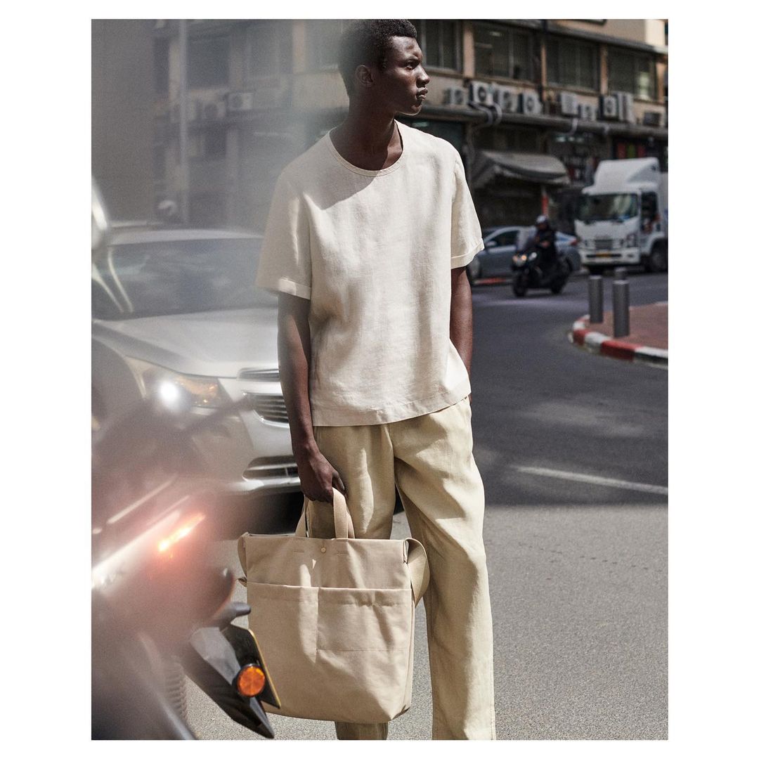A dark-skinned male model in a beige t-shirt and slacks stands on a city street holding a beige tote bag with cars and buildings behind him.