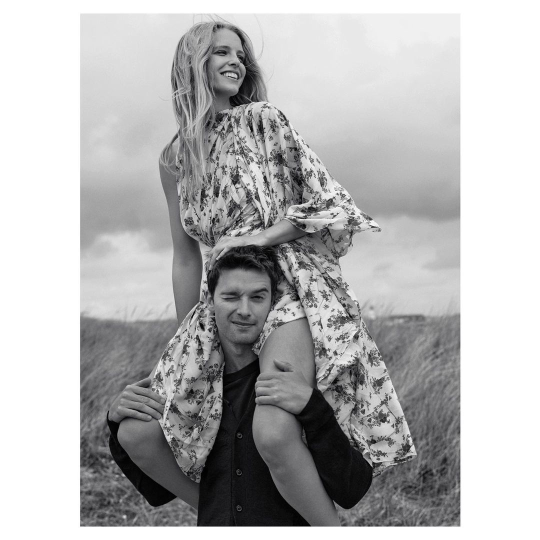 A smiling woman in a floral dress sits on a man's shoulders against a cloudy sky and grassy field in a black and white outdoor portrait.