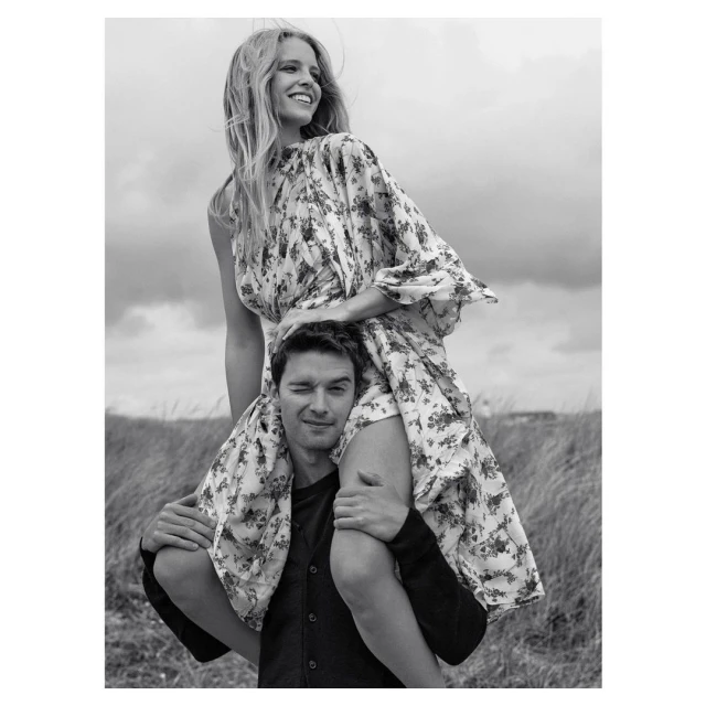 A smiling woman in a floral dress sits on a man's shoulders against a cloudy sky and grassy field in a black and white outdoor portrait.
