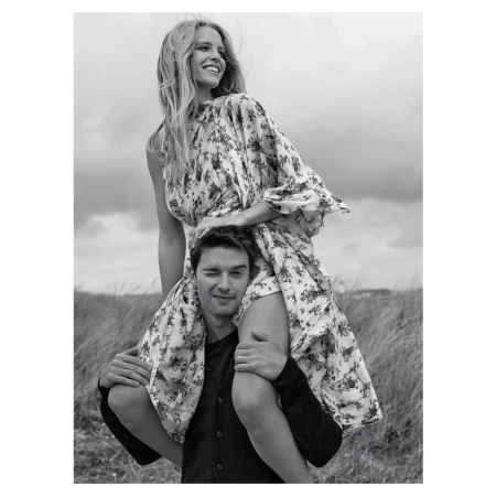 A smiling woman in a floral dress sits on a man's shoulders against a cloudy sky and grassy field in a black and white outdoor portrait.
