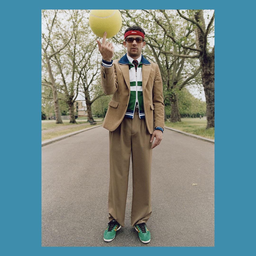 Man in tan suit and green sneakers balances a tennis ball on one finger in an outdoor scene with trees and an asphalt path.