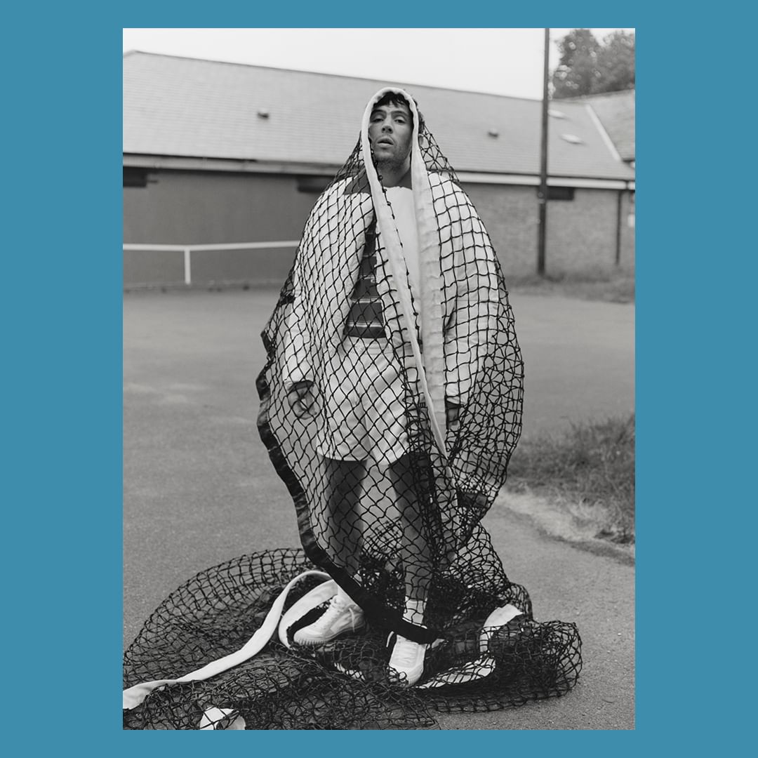 Monochrome portrait of a man draped in a fishnet over a white outfit, standing on an outdoor tennis court with a building in the background.