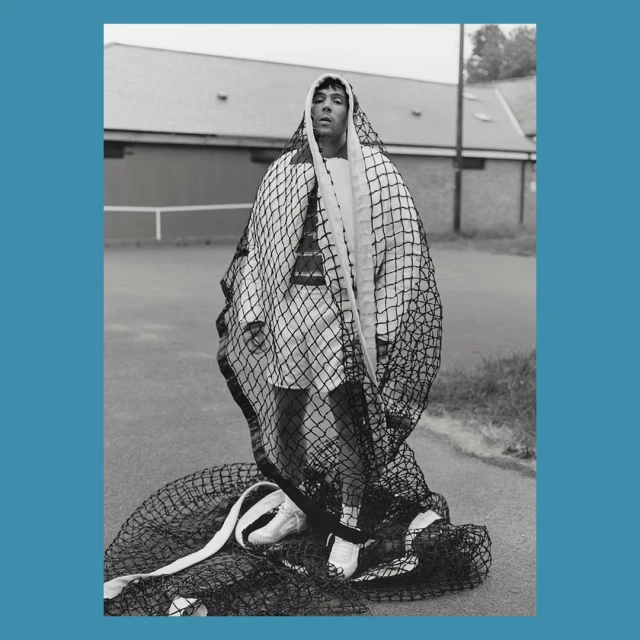 Monochrome portrait of a man draped in a fishnet over a white outfit, standing on an outdoor tennis court with a building in the background.