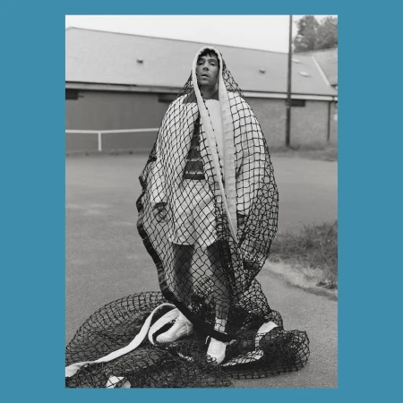 Monochrome portrait of a man draped in a fishnet over a white outfit, standing on an outdoor tennis court with a building in the background.