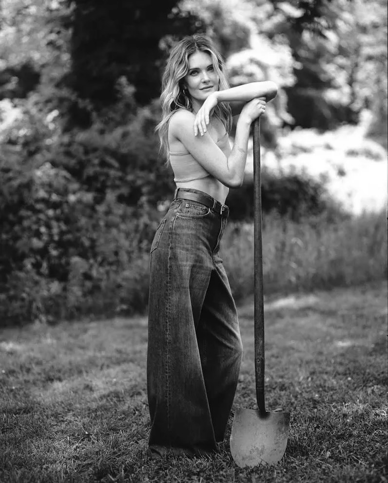 Black and white portrait of a woman leaning on a shovel, wearing jeans, a top, and jewelry standing in a grassy outdoor setting.