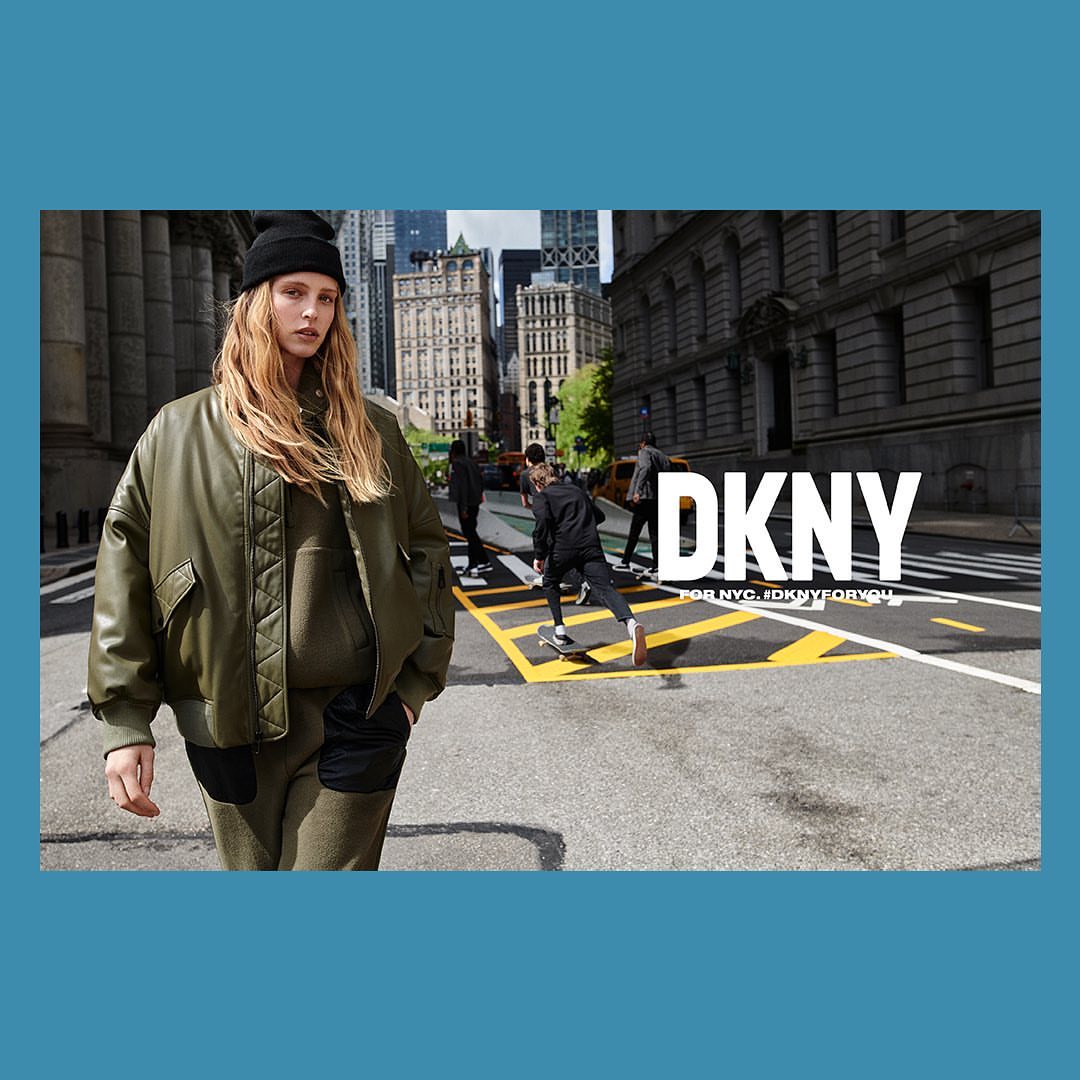 Fashion image featuring a blonde adult female in an olive green jacket and beanie, walking on a city street with skateboarders in the background.
