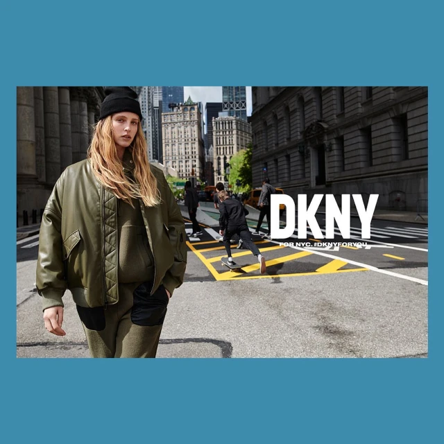 Fashion image featuring a blonde adult female in an olive green jacket and beanie, walking on a city street with skateboarders in the background.