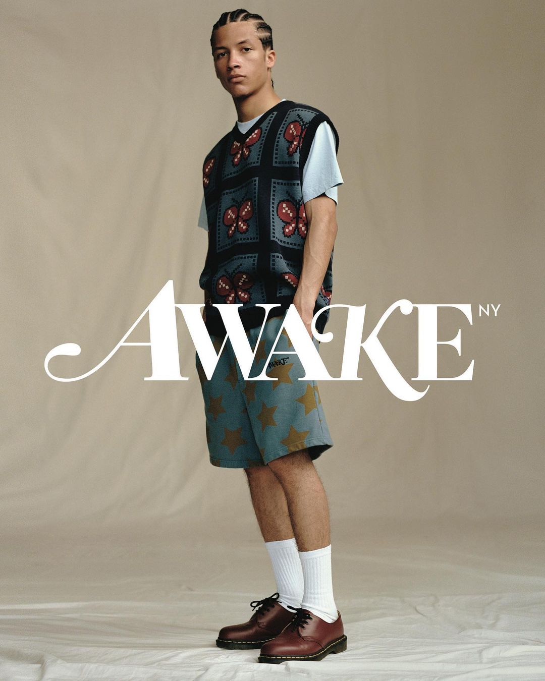 Male model wearing a butterfly sweater vest, star shorts, white socks, and dress shoes in a studio setting with the brand logo.