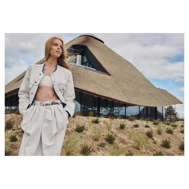 Woman poses in a denim jacket and pants near a large, modern thatched-roof house on a grassy hill under an overcast sky.
