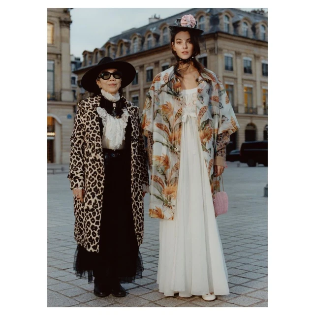 Two women in fashionable coats pose outdoors. Woman in leopard print coat and sunglasses stands next to woman in floral print coat and hat.