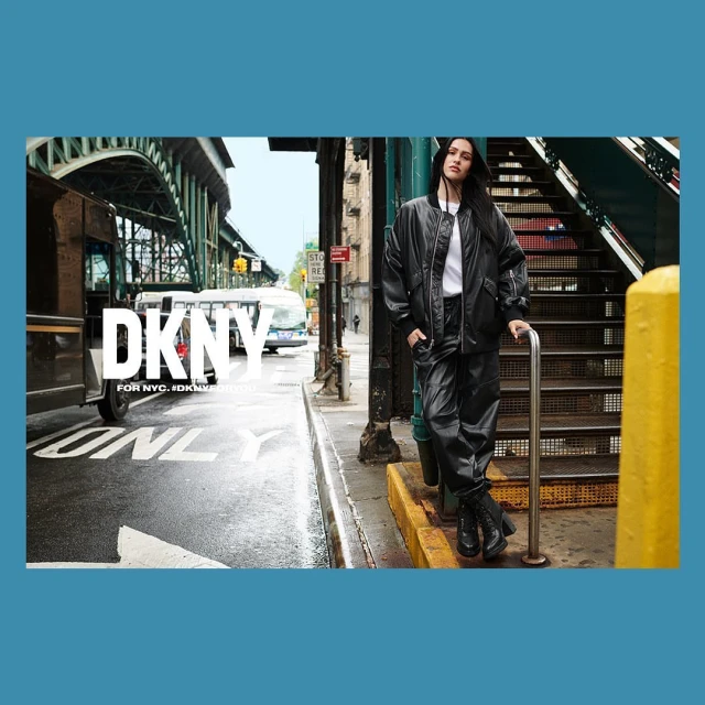 Fashion shot of a woman in a black jacket and pants posing on an urban street next to steps, with a bus and an overpass in the background.
