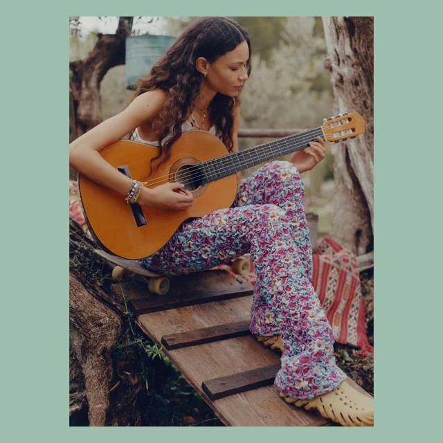 Young adult woman with long dark hair, wearing floral pants and clogs, sits on a skateboard while playing a brown acoustic guitar.