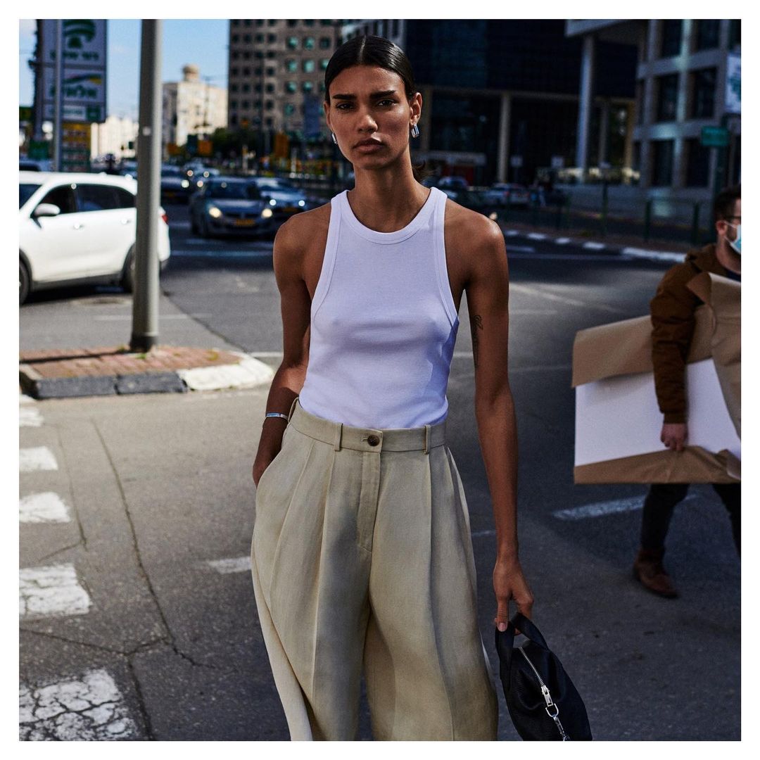 A woman in white top and khaki pants stands in a city street with cars and buildings in the background on a sunny day.