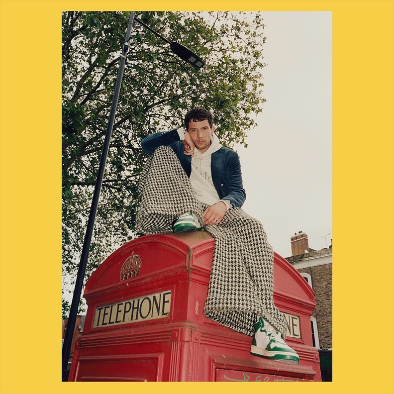 A young adult male sits on a red telephone booth wearing a blazer, hoodie, and patterned pants in an outdoor urban setting with trees.