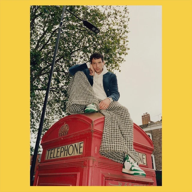 A young adult male sits on a red telephone booth wearing a blazer, hoodie, and patterned pants in an outdoor urban setting with trees.