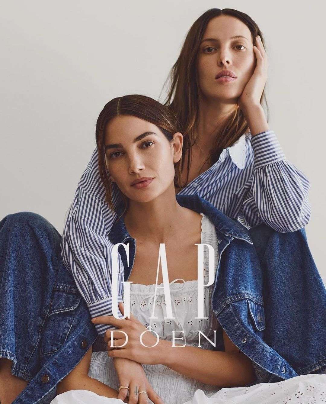 Two women in a fashion advertisement, one with hand on face, wearing denim jacket and white dress, against a plain neutral background.