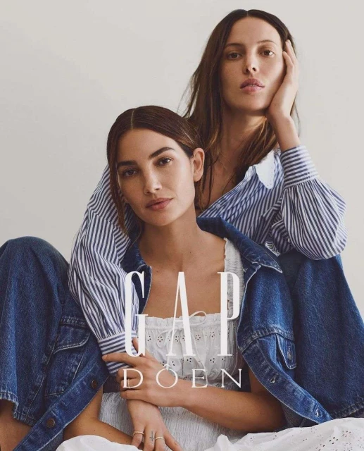 Two women in a fashion advertisement, one with hand on face, wearing denim jacket and white dress, against a plain neutral background.