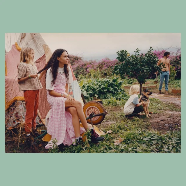 Family scene outdoors with two children and woman in a garden with tents, lush greenery, and a dog, under an overcast sky.