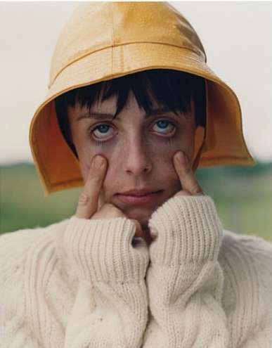 Close-up portrait of a young adult in a yellow bucket hat and white cable knit sweater, outdoors with green field background.