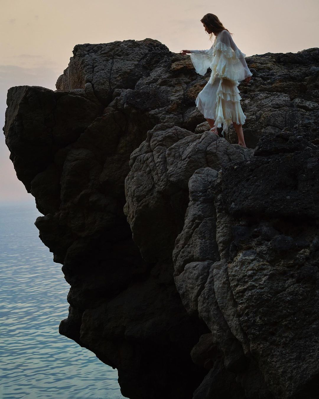 Woman in white dress on a rocky cliff overlooking the ocean at dusk. Dramatic, natural outdoor setting with tranquil water.