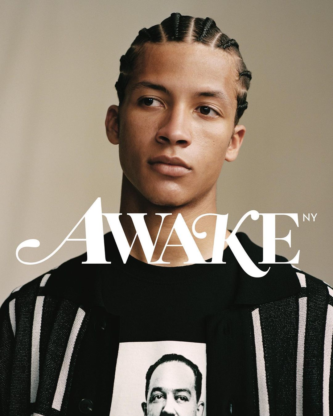 Portrait of a young adult male with braided hair wearing a black graphic tee with an image and a black and white striped jacket.