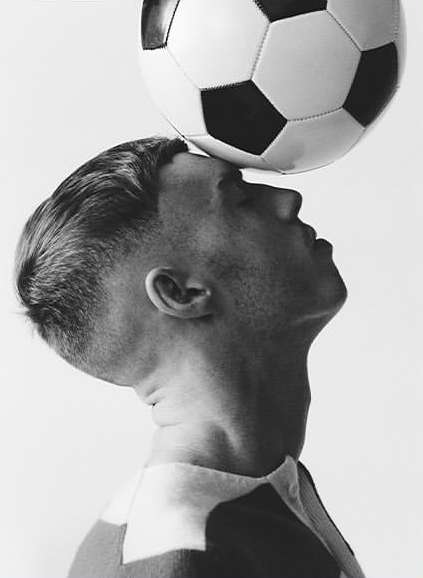 Profile of a young man balancing a classic black and white soccer ball on his forehead against a plain white background.