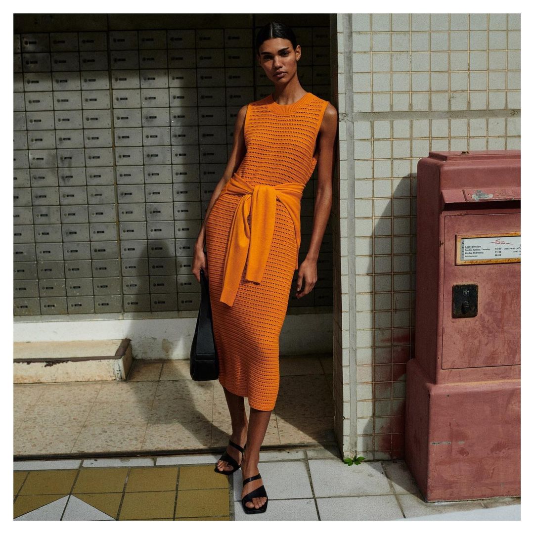 Woman in an orange sleeveless dress and black sandals stands in front of a tile wall with a mailbox in an outdoor setting.