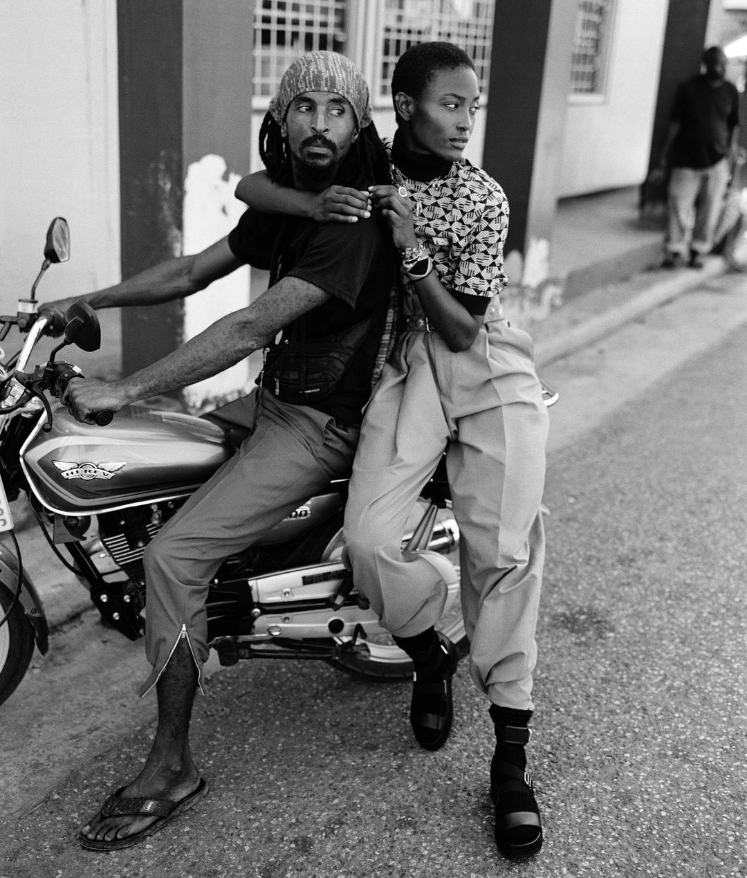 Black and white shot of a man on a motorcycle with a woman behind him, showcasing fashion in an urban setting with natural lighting.