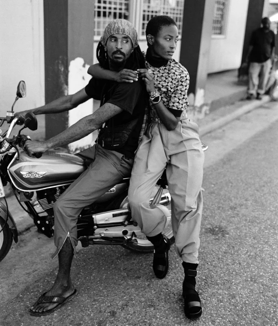Black and white shot of a man on a motorcycle with a woman behind him, showcasing fashion in an urban setting with natural lighting.