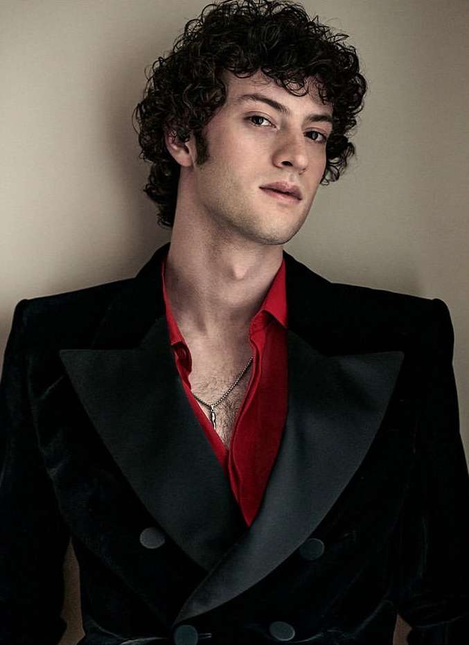 Portrait of a fair-skinned adult male with curly dark hair, wearing a black velvet blazer over a red collared shirt, indoors.