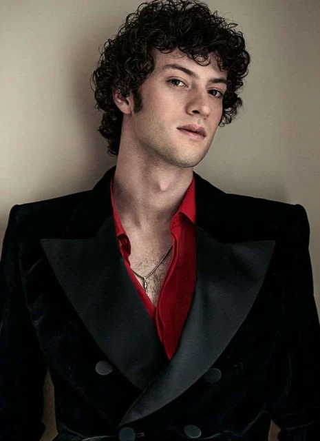 Portrait of a fair-skinned adult male with curly dark hair, wearing a black velvet blazer over a red collared shirt, indoors.