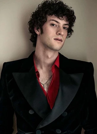 Portrait of a fair-skinned adult male with curly dark hair, wearing a black velvet blazer over a red collared shirt, indoors.