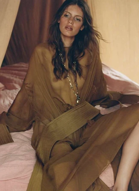 A young woman in a sheer, belted brown outfit poses while seated indoors on a light pink bed, bathed in soft light.