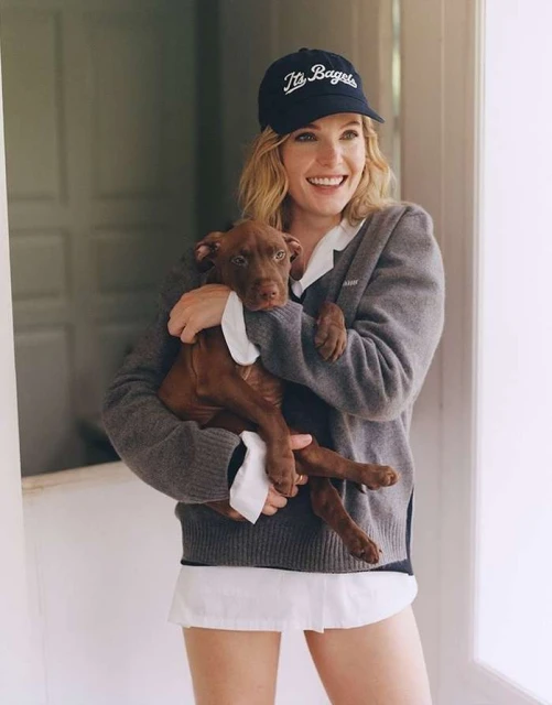 A woman smiles warmly, holding a brown puppy while standing inside a white doorway. The woman wears a baseball cap, sweater and a white top.