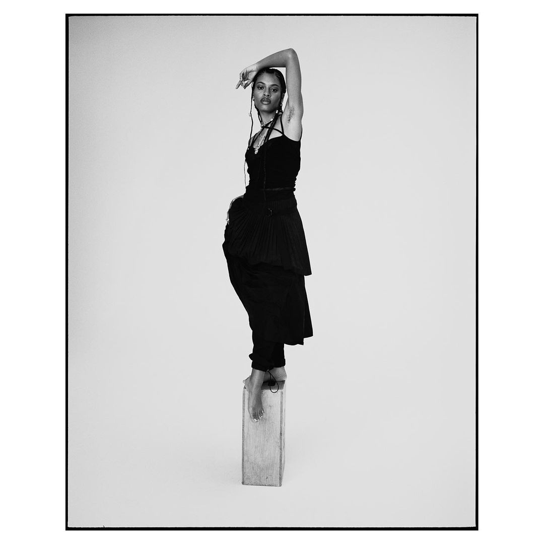 Woman in black dress poses on wooden block against a light grey background. Black and white studio fashion shot, full length.