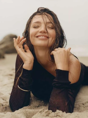 Smiling woman with wet hair lying on a sandy beach wearing a dark long-sleeve top