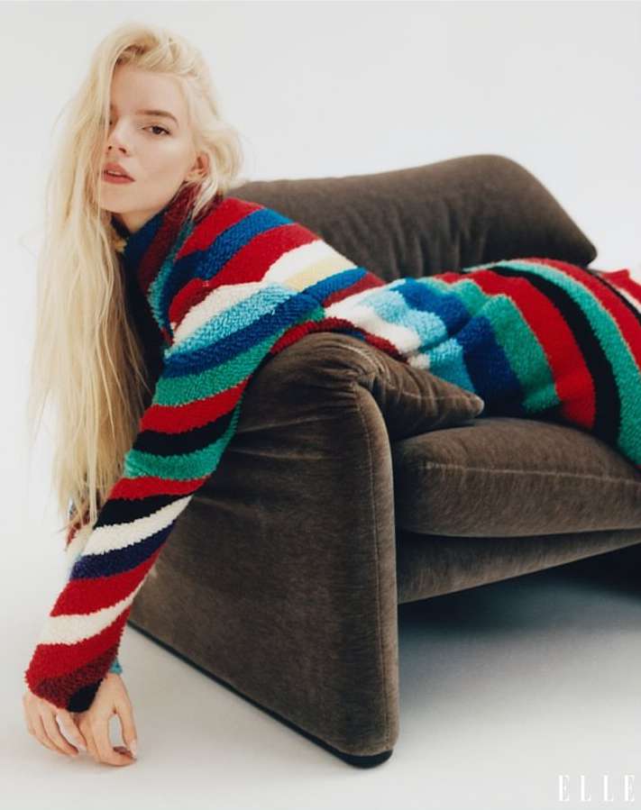 Attractive blonde woman in colorful striped sweater poses on brown couch in a studio setting with soft, diffused light for Elle magazine.