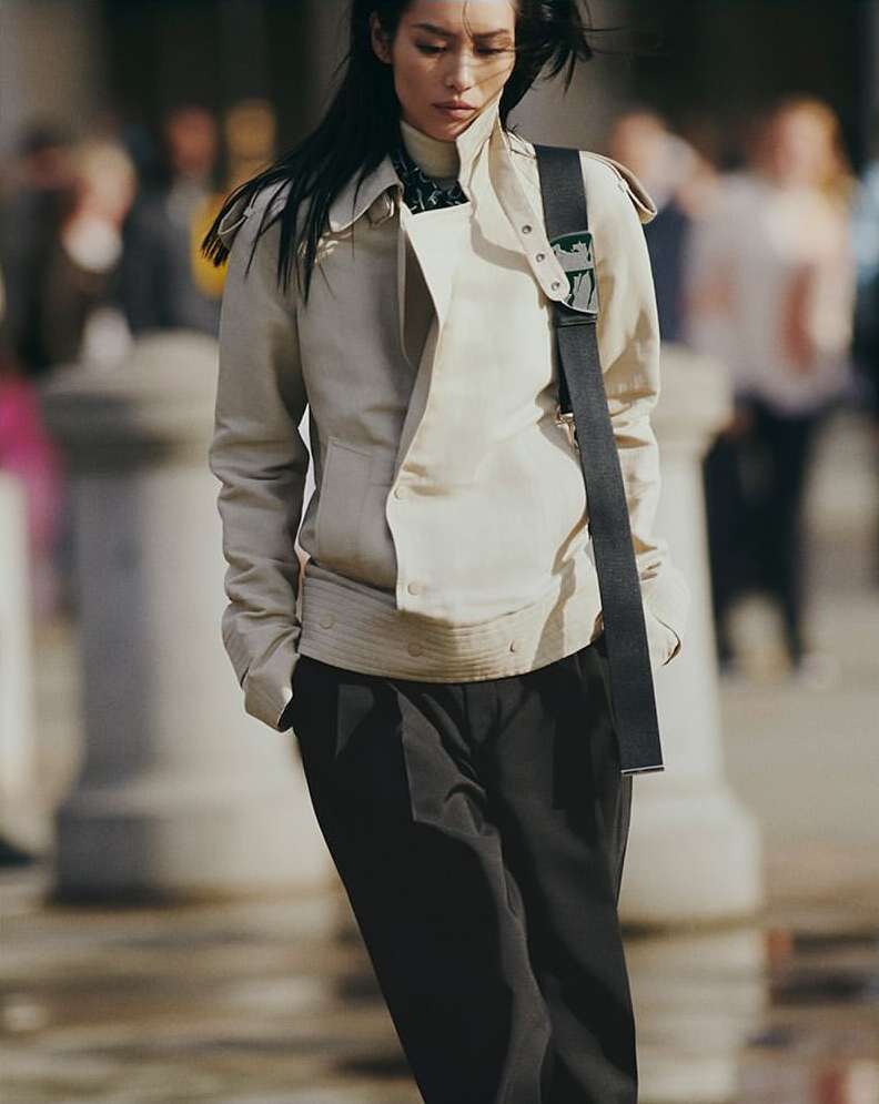 An Asian woman is walking outdoors, wearing a beige jacket, black pants, and shoulder bag, in a medium shot with a blurred background.