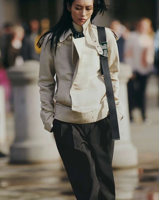 An Asian woman is walking outdoors, wearing a beige jacket, black pants, and shoulder bag, in a medium shot with a blurred background.