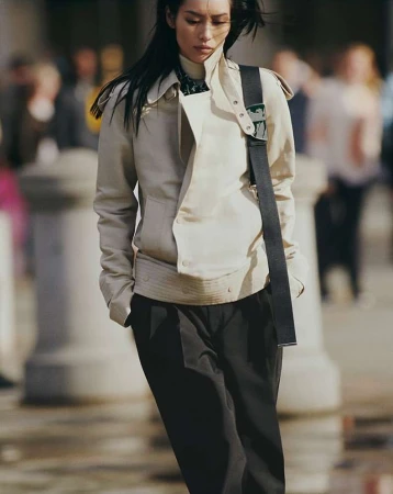 An Asian woman is walking outdoors, wearing a beige jacket, black pants, and shoulder bag, in a medium shot with a blurred background.