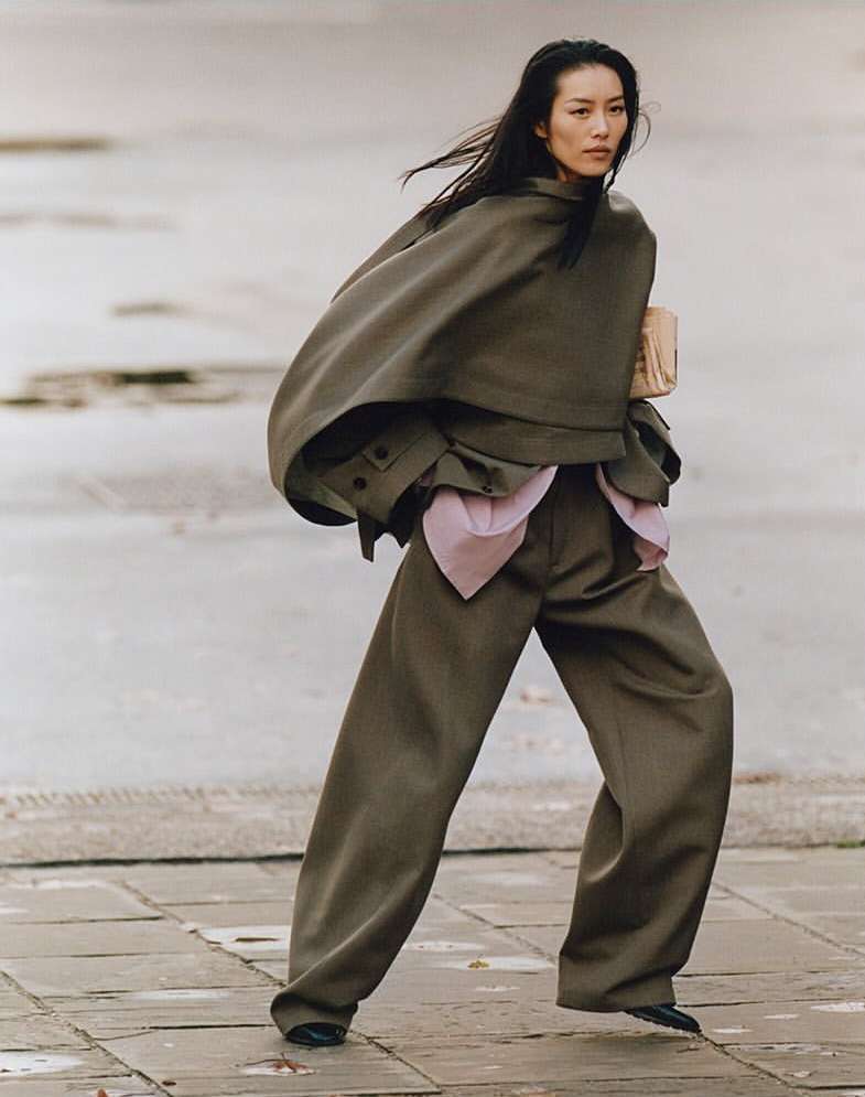 A woman wearing an olive-green cape and pantsuit poses on a stone surface with natural lighting, showcasing fashion and style.