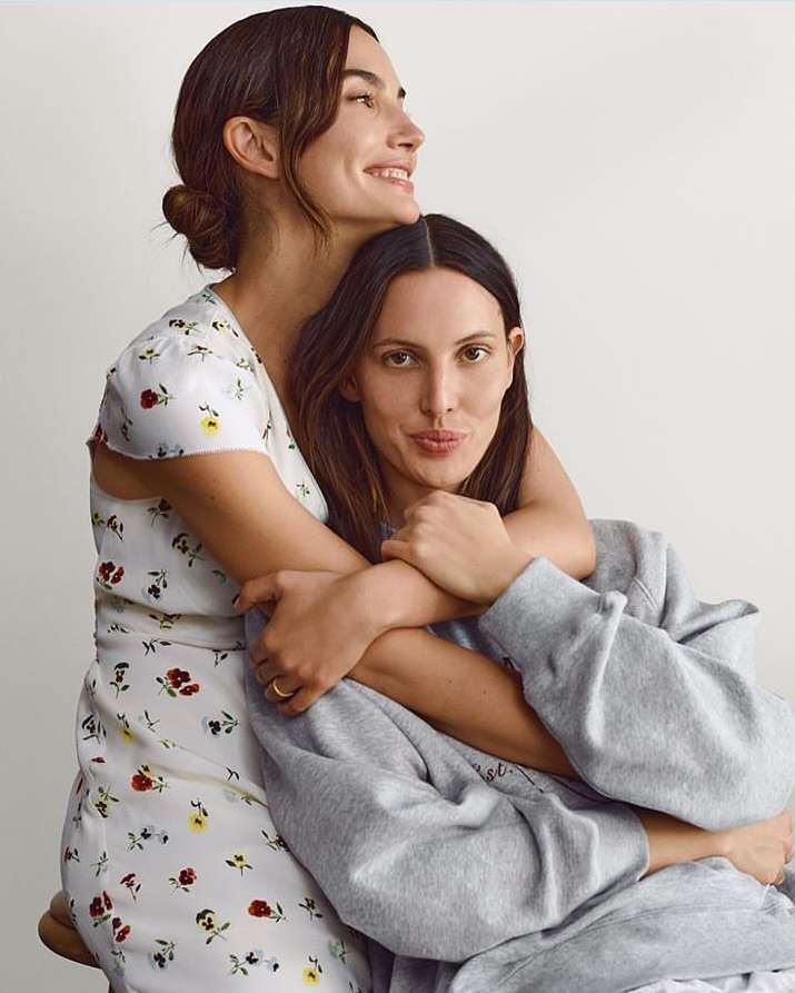 Two adult females, embracing indoors with natural light. One woman wears a floral dress, the other a grey sweatshirt. Both have dark hair.