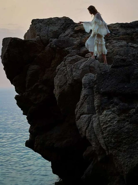 Woman in white dress stands on rocky cliff edge overlooking ocean. Sunset sky and dramatic rock formations create a scenic view.