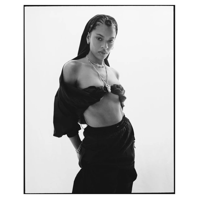 A striking black and white portrait of an adult female with cornrow braids, wearing a scarf, necklace, and draped fabric top in a photo studio.