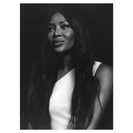 Black and white portrait of Naomi Campbell with long dark hair wearing a white sleeveless top against a dark background