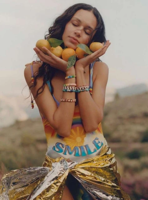 Woman poses outdoors holding oranges and wearing patterned top and gold wrap. She has beaded bracelets and wavy brown hair.