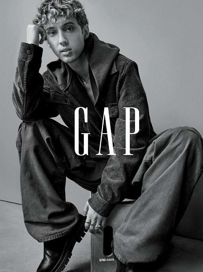 Monochrome portrait of a young adult male model in casual apparel sitting on a box with the logo 'GAP' dominating the foreground.